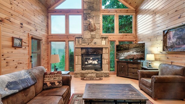 A cozy cabin interior with a stone fireplace, wooden walls, and a large window overlooking greenery.
