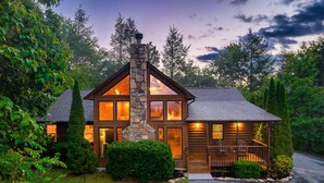 A log cabin with a stone chimney, large windows, and a porch with chairs.