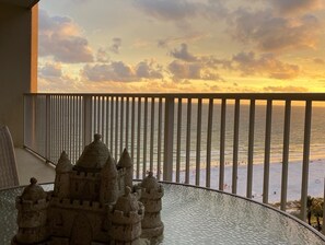 A balcony with a view of the sea at sunset, a glass table, and a sandcastle model.