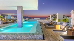 Piscina infinita en la azotea con vistas al mar al atardecer, sillones de descanso y sombrillas.
