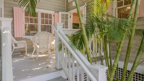 A porch with white wicker chairs, a small table, and a staircase leading to a balcony with bamboo plants. - One Bedroom Deck Villa Steps from Duval! (Key West)