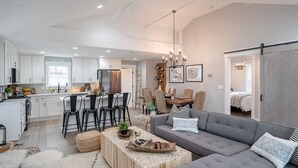 Una cocina moderna con armarios blancos, un comedor con candelabro y una sala de estar con sofá gris y una mesita de centro.