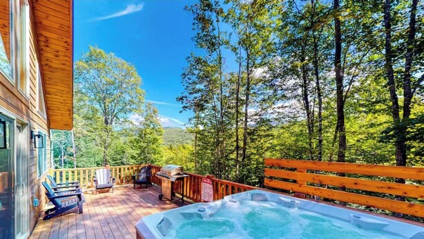 A wooden deck with a hot tub, outdoor seating, and a view of a forested area. - Gorgeous dog-friendly cabin with great view, deck, hot tub, & gas fireplace (Kingfield)