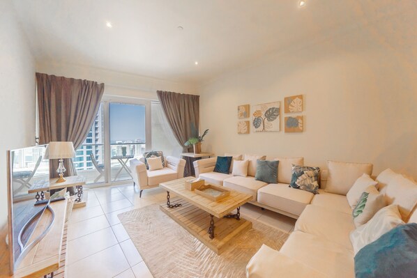 A modern living room with a sofa, coffee table, and a TV. There is a balcony with curtains and a view of buildings.