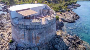 Une structure cylindrique en pierre, dotée d’un toit plat et d’un espace patio aménagé avec des chaises longues et une table.