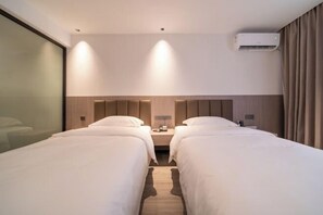 A hotel room with two beds, a bedside table with a lamp, and an air conditioning unit on the wall.