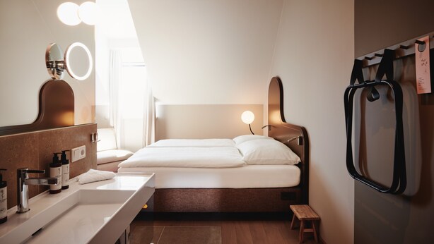 Una habitación de hotel moderna con una cama grande, un tocador con espejo e iluminación, un taburete pequeño y un perchero de pared.