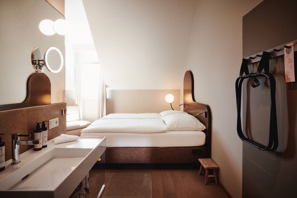 Quarto de hotel moderno com uma cama grande, uma mesa de toucador com espelho e iluminação, um banquinho pequeno e uma estante fixada na parede.