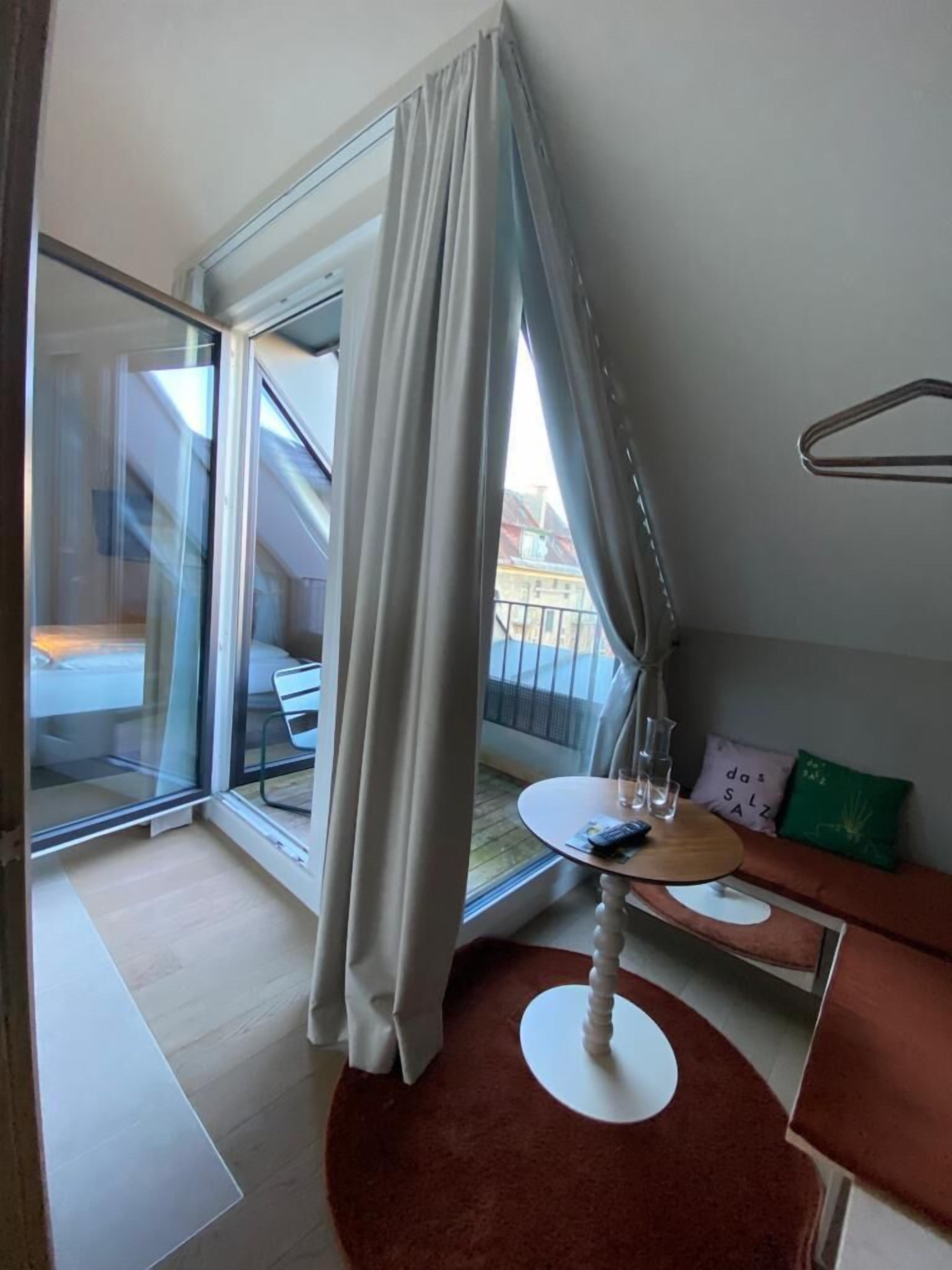 Een moderne kamer met een glazen deur naar een balkon, een ronde tafel met een stoel, en een bank met kussens.