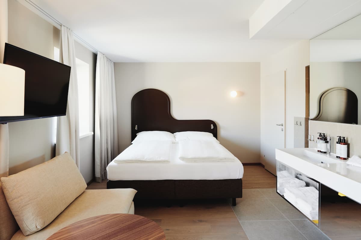Quarto de hotel moderno com uma cama grande, mesa de cabeceira, televisão de tela plana, toucador embutido com espelho e sofá.