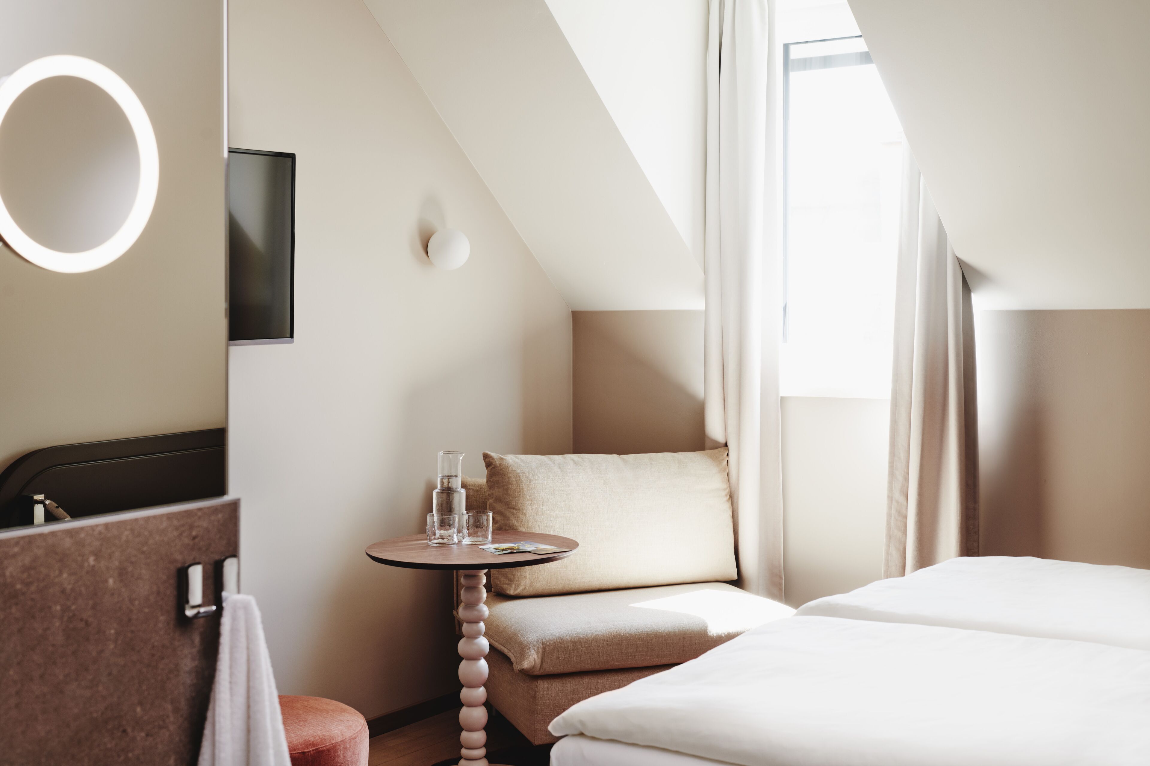 Une chambre d’hôtel moderne, équipée d’un lit, d’une petite table ronde avec des verres, d’un canapé et d’une grande fenêtre avec des rideaux.