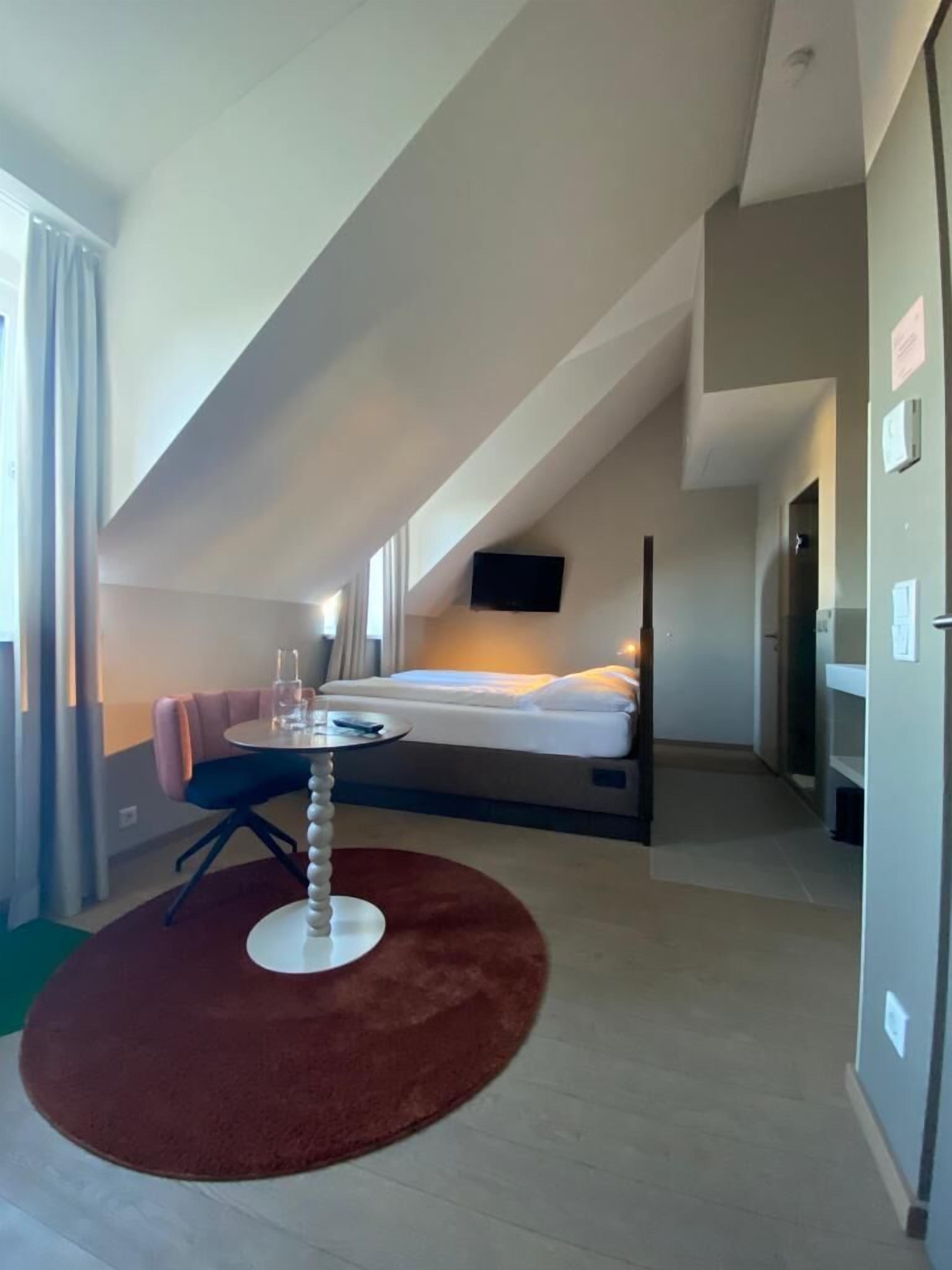 A modern hotel room with a sloped ceiling, a bed, a small round table, and a red rug.