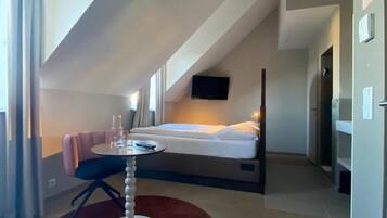 A modern hotel room with a sloped ceiling, a bed, a small round table, and a red rug.