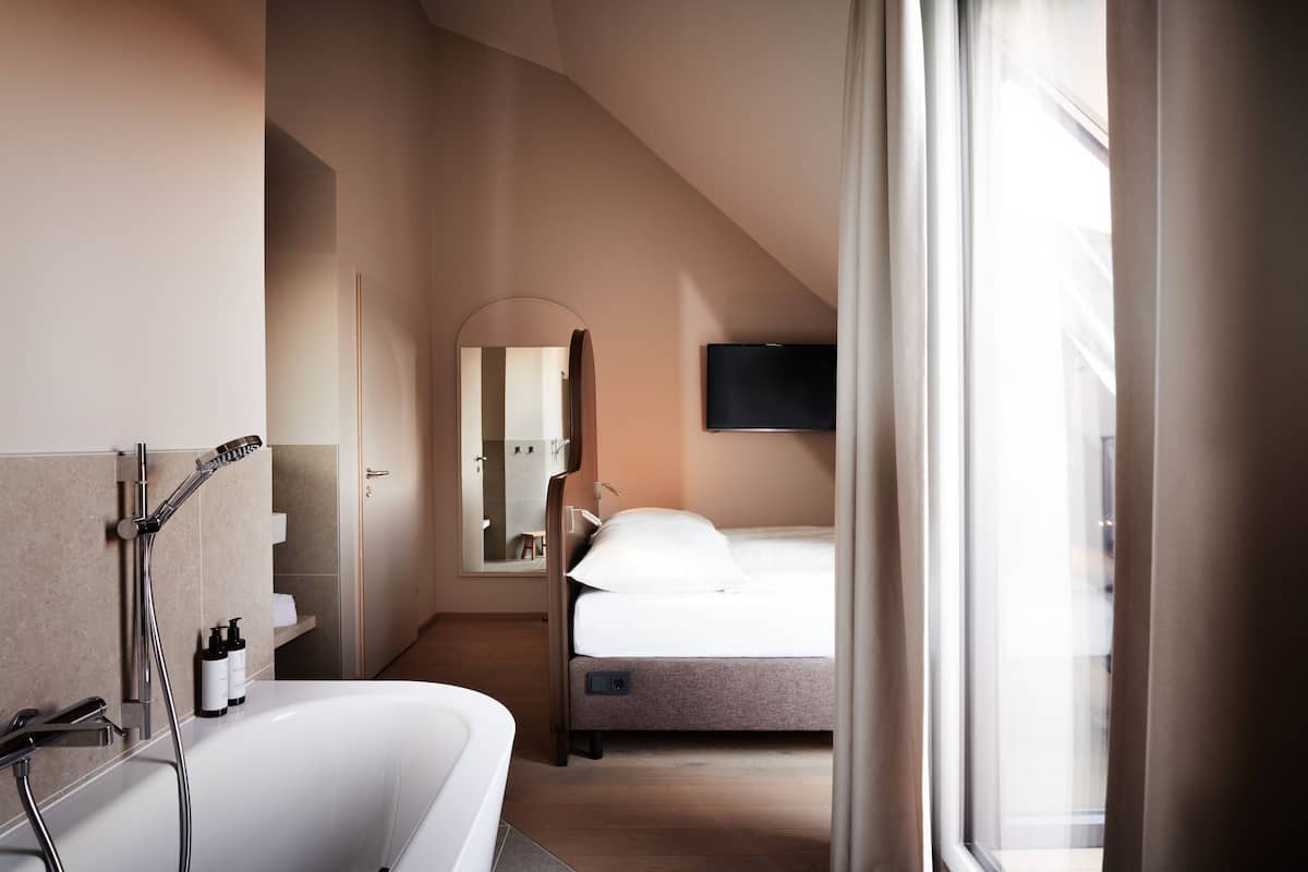 Quarto de hotel moderno com uma janela grande, uma banheira, uma cama e um espelho.