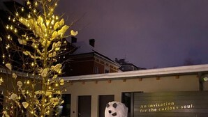 A snowman sculpture in a snowy outdoor area with tables and chairs, and a building with a sign that reads "An invitation for the curious soul."