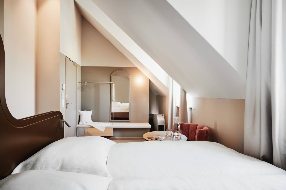 Quarto de hotel moderno com uma cama grande, uma mesa com cadeira, uma pequena mesa com copos e um banheiro visível através de uma porta aberta.