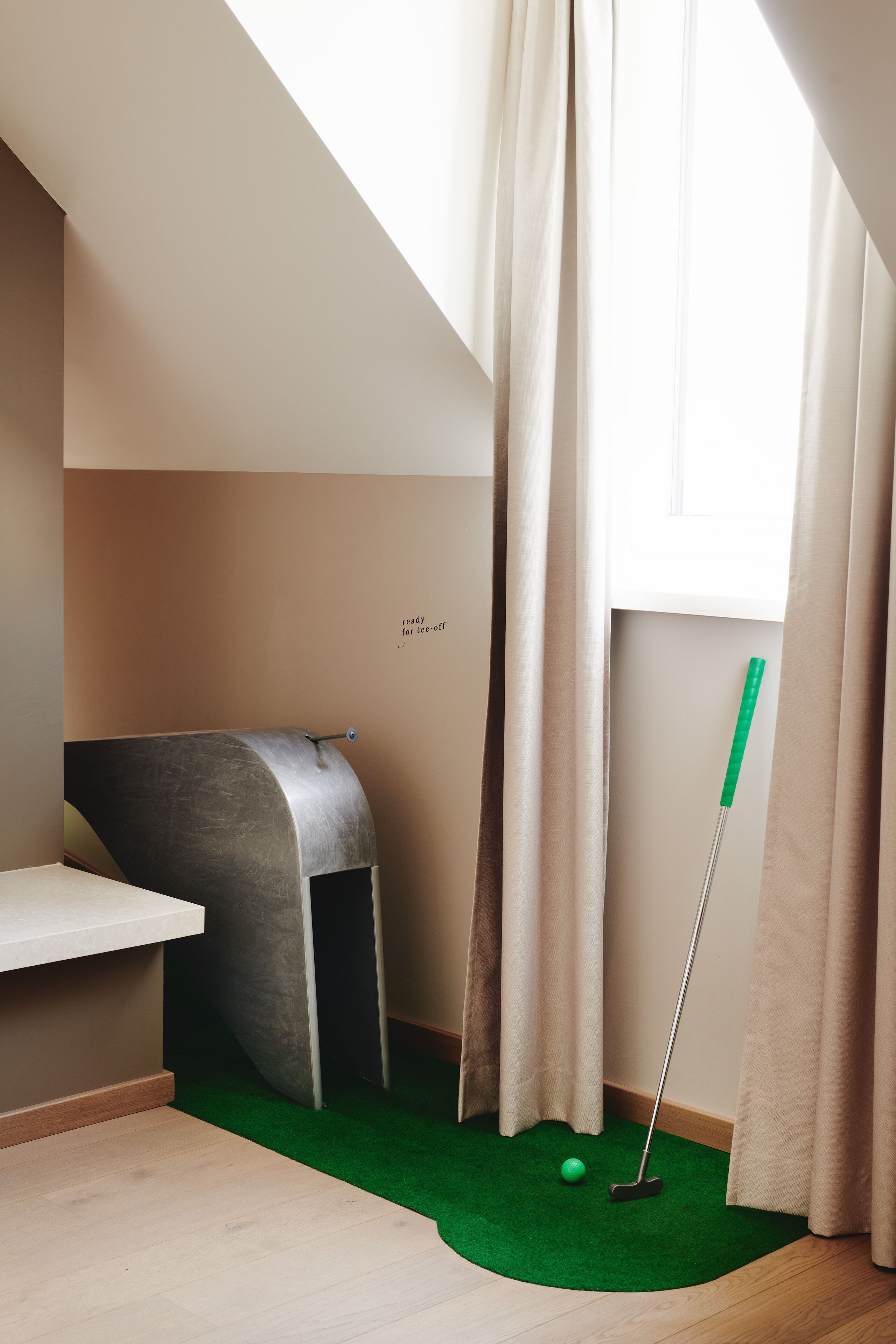 Ein Zimmer mit einem begrünten Rasenbereich, einer Metallbank, einem Fenster mit Vorhängen und einem Schild mit der Aufschrift 'bereit für den Tee-Off'.