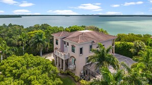 A two-story house with a red-tiled roof, surrounded by lush greenery and a large body of water. - Huge Estate, Heated Pool, Water View, King beds, Sleeps 20 (Marco Island)
