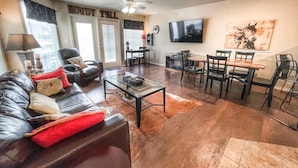 A spacious living room with a leather couch, a glass coffee table, and a dining area with a TV and artwork on the wall. - Poolside Perch | Sleeps 6 | Pet-Friendly (New Braunfels)