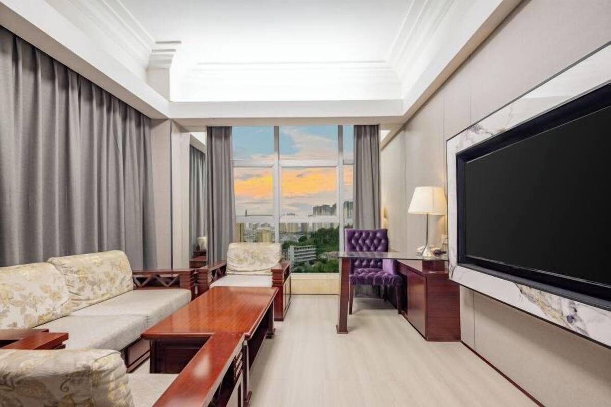 Habitación de hotel con televisor grande, sofá, mesa de comedor y vistas a la ciudad.