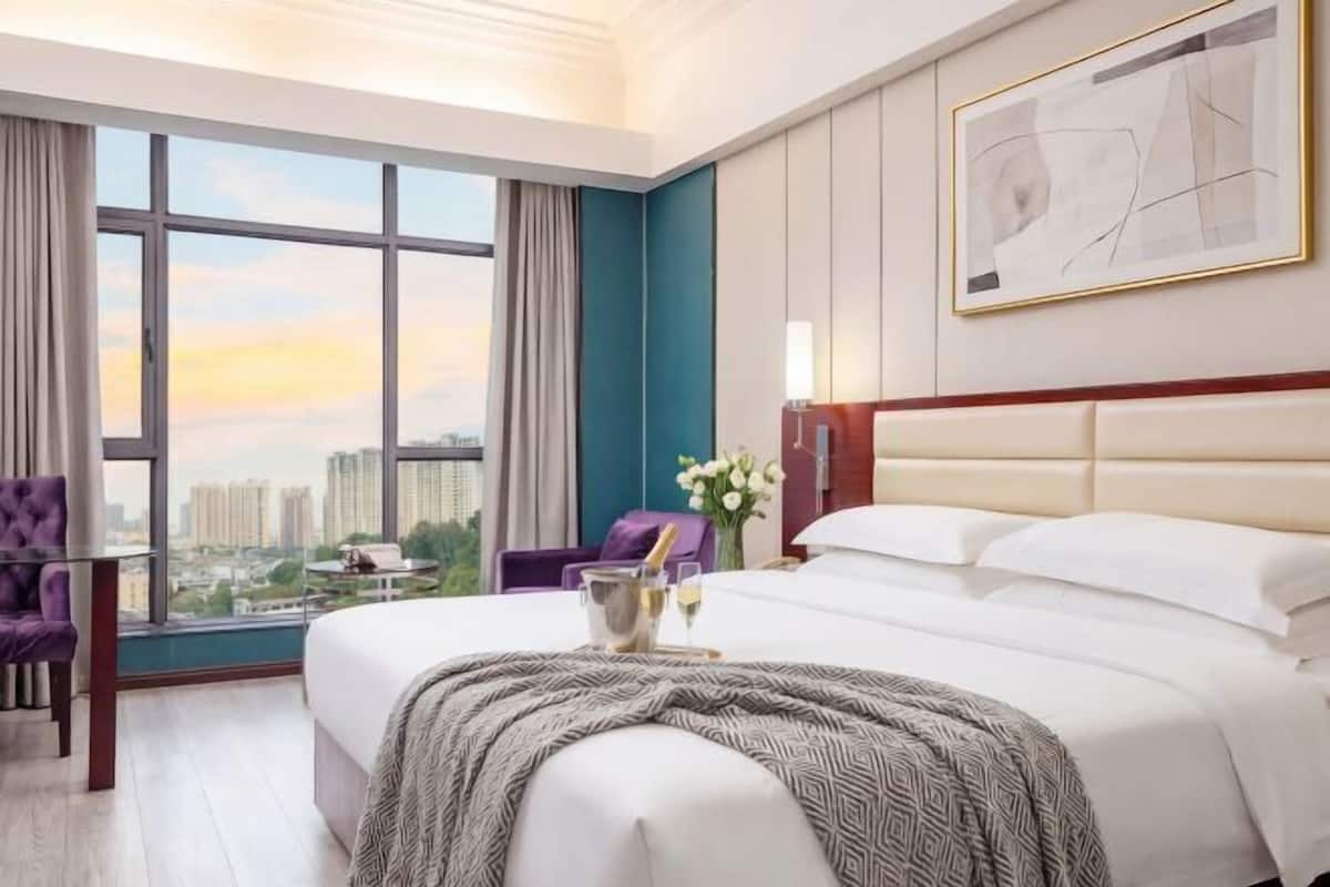Un cuarto de hotel con una cama grande, vista a la ciudad, una silla morada y una mesa con un jarrón de flores.