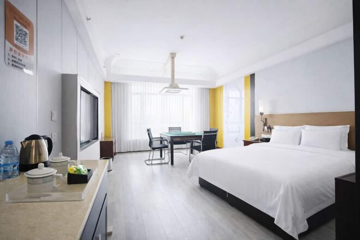 Habitación de hotel con cama, escritorio con silla, mesa, hervidor, botella de agua y ventana con cortinas.
