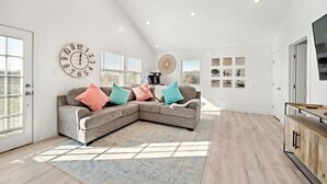A spacious living room with a large sectional sofa, a round wall clock, and a large window with blinds. - Cozy Home Near Seawall with Private Pool (Galveston)
