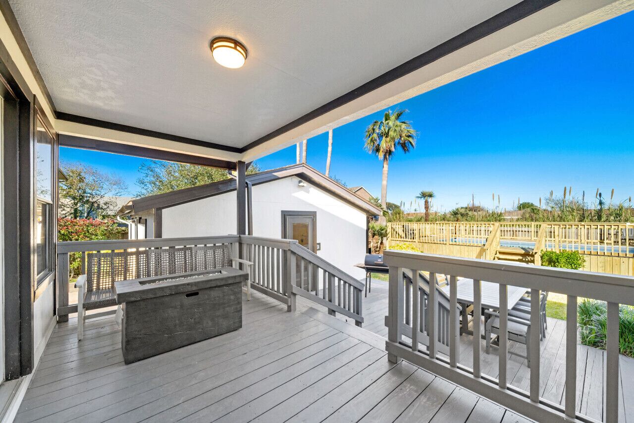 A covered outdoor deck with a built-in bar area, a staircase, and a view of a landscaped garden and palm tree.