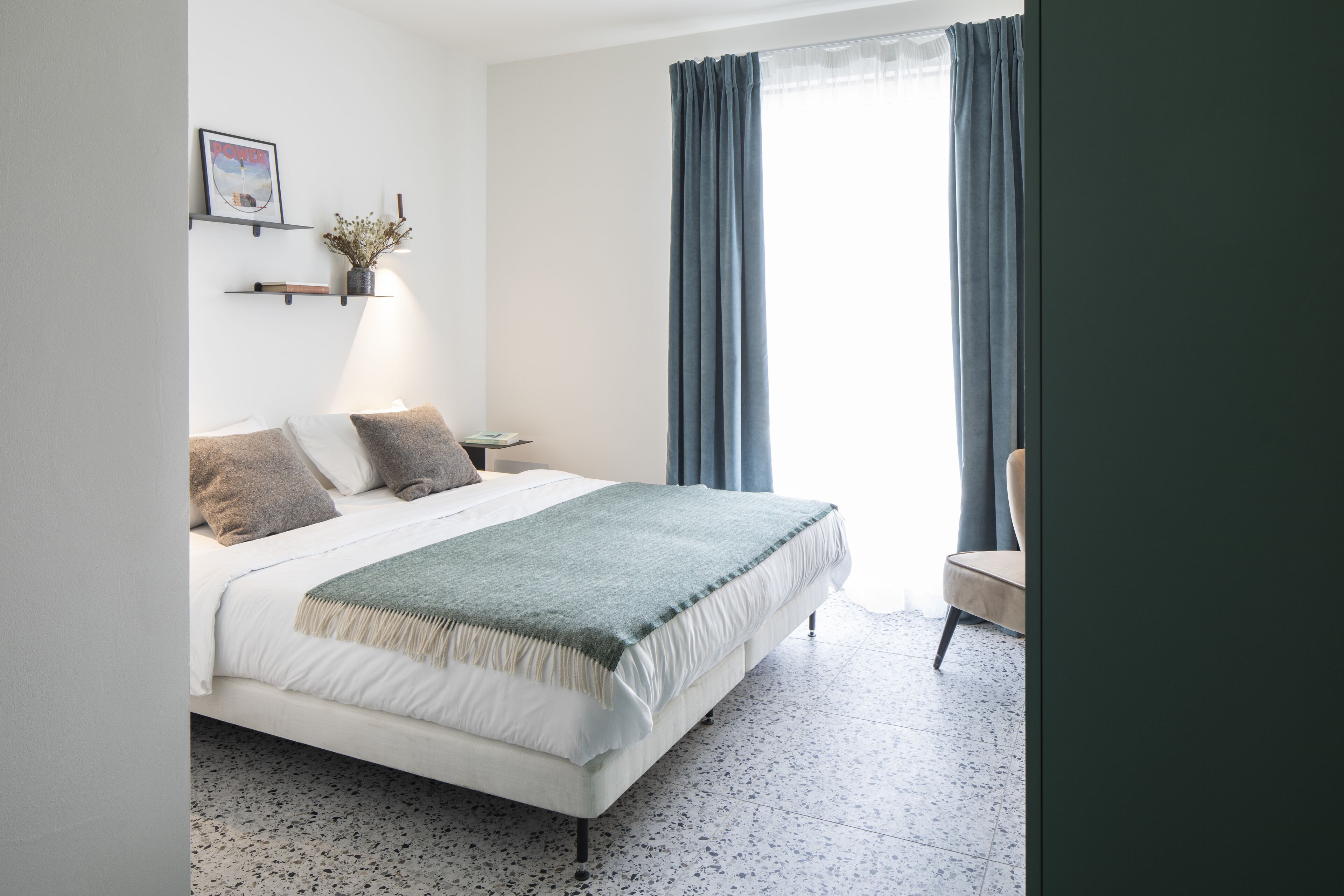 A bedroom with a bed, a chair, a nightstand, and a wall-mounted shelf with a plant and candles.
