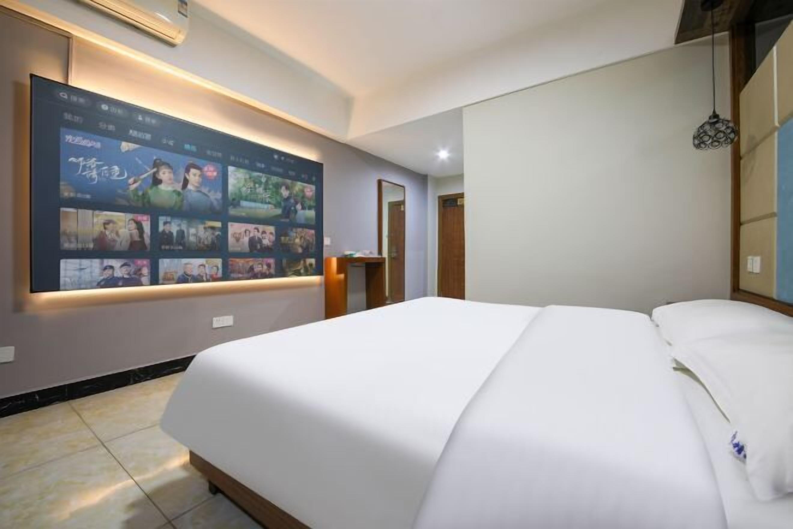 A hotel room with a large wall-mounted TV displaying various shows, a neatly made bed, and a small desk with a lamp.