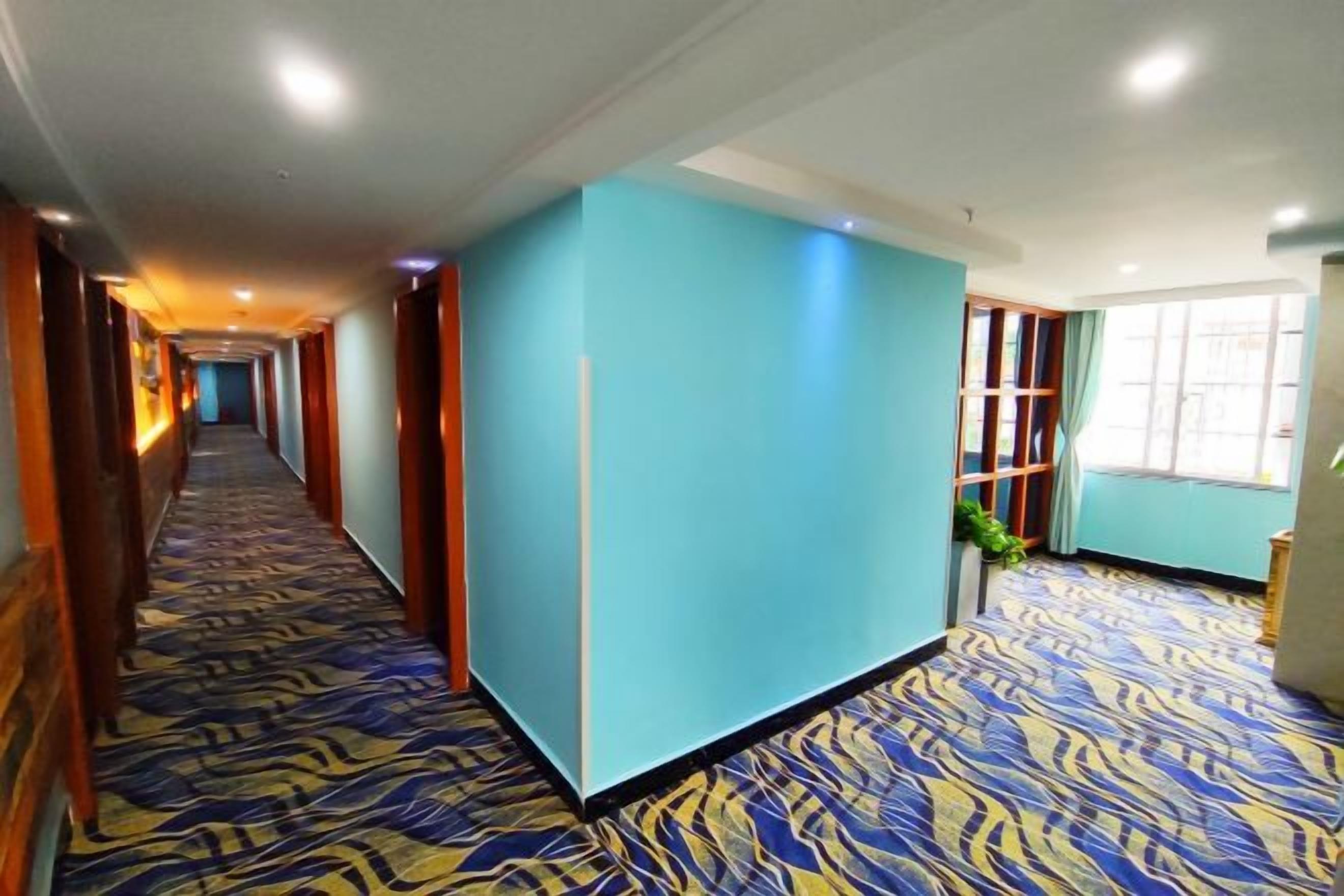 Ein Hotelgang mit einem gemusterten Teppich, türkisfarbenen Wänden und einem Fenster auf der rechten Seite.