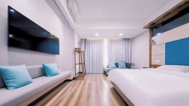 Habitación de hotel con piso de madera, sofá con cojines azules, cama con ropa blanca, televisor de pantalla plana montado en la pared y ventana con cortinas.