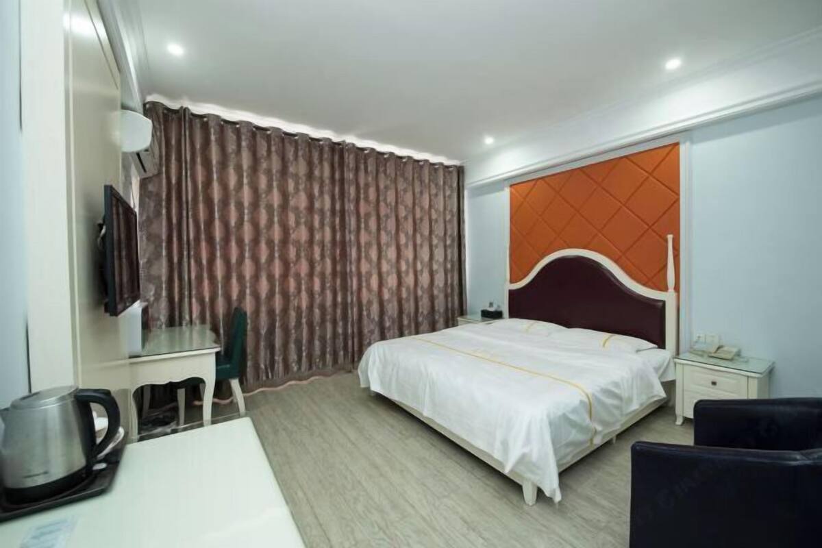 A hotel room with a bed, a desk with a kettle, a chair, a nightstand, and a television.