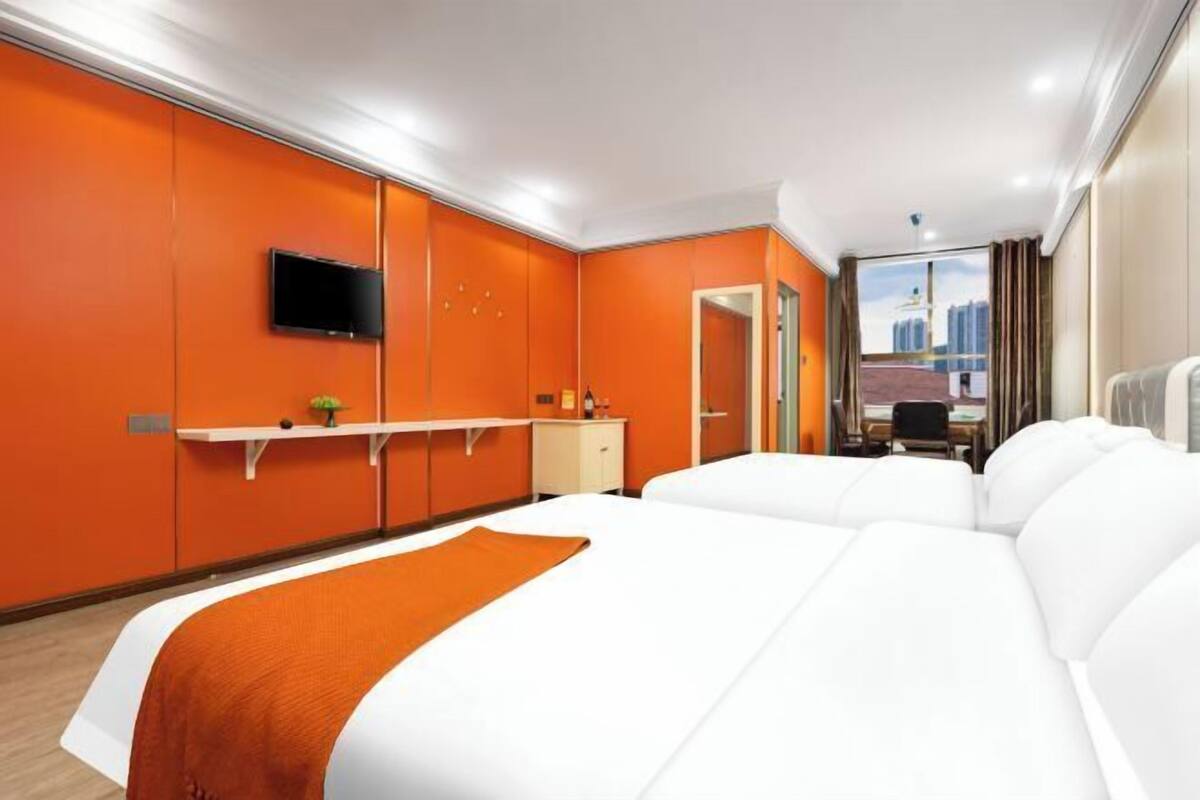 A hotel room with a large bed, a TV mounted on the wall, a desk with a chair, and a view of the cityscape through the window.