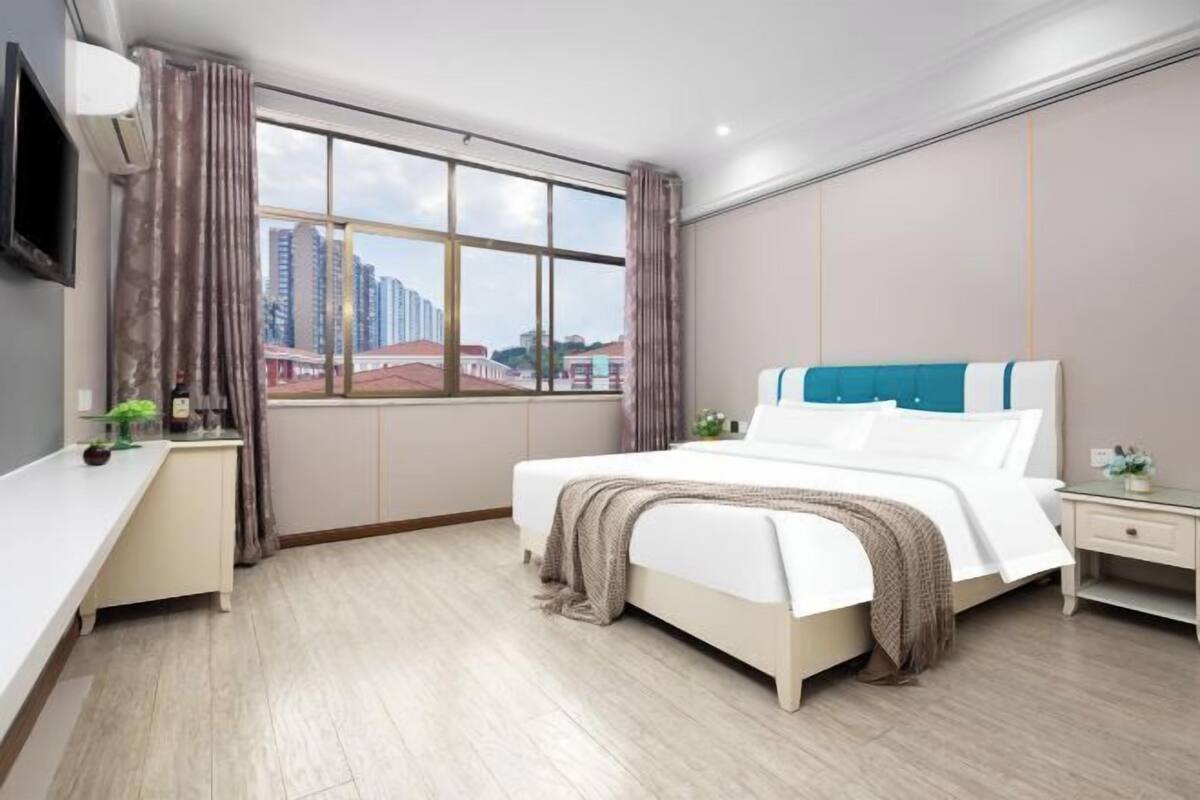A modern hotel room with a large window, a bed, a bedside table, and a desk.