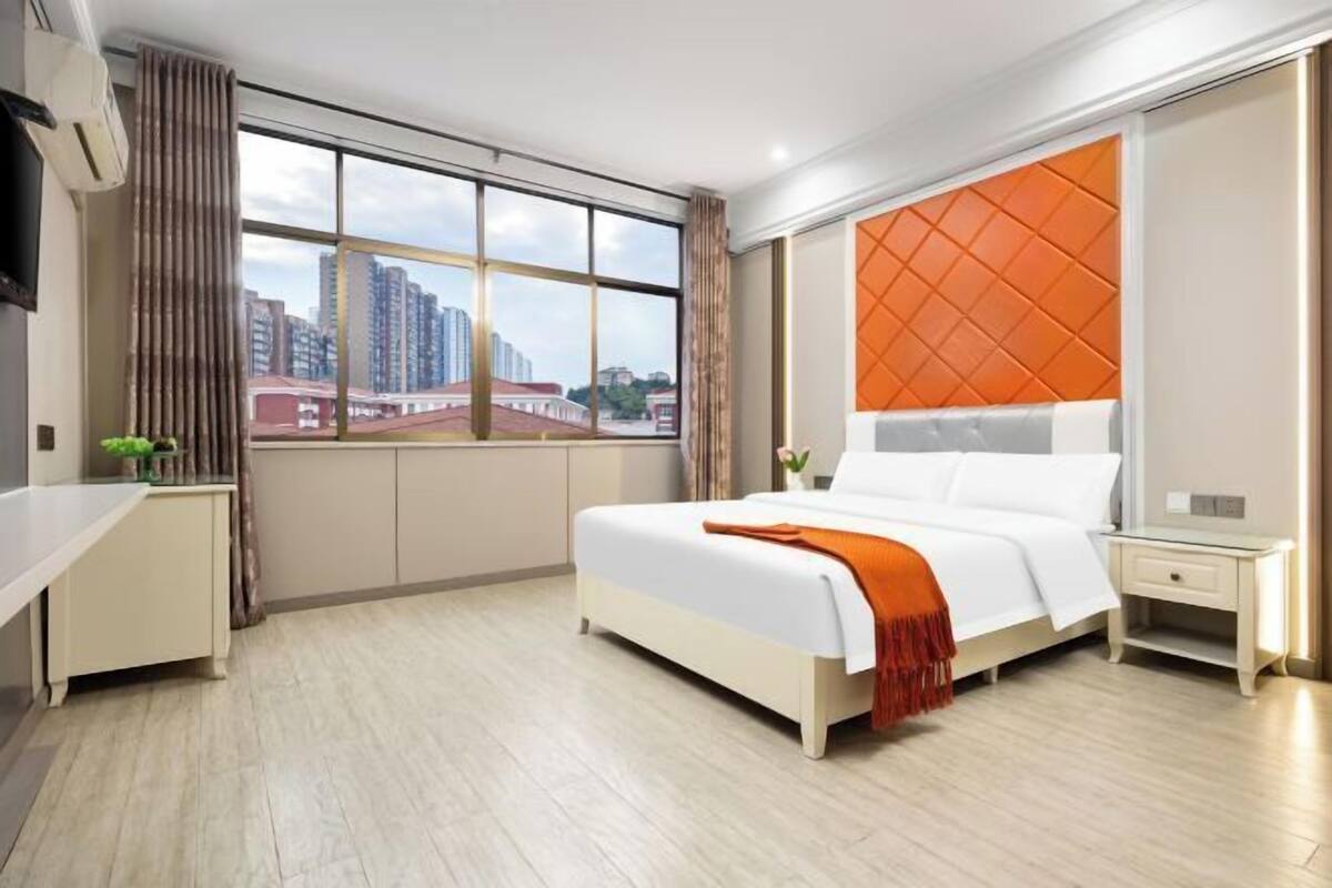 A modern hotel room with a large bed, a desk, a nightstand, and a window with a city view.
