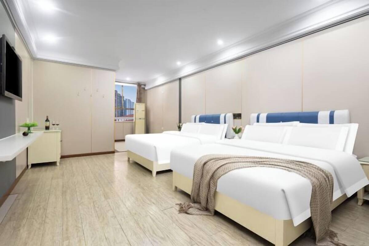 A modern hotel room with two beds, a flat-screen TV, a small bedside table, and a window with a cityscape view.