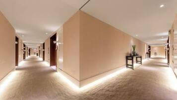 A hotel corridor with a carpeted floor, wall-mounted lights, and a reception desk.