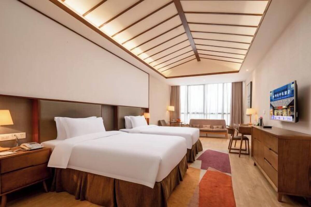 Ein Hotelzimmer mit zwei Betten, einem Schreibtisch, einem Fernseher und einem großen Fenster.