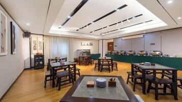 A hotel dining area with multiple tables and chairs, a kitchen in the background, and a high ceiling with recessed lighting.