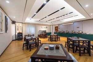 A hotel dining area with multiple tables and chairs, a kitchen in the background, and a high ceiling with recessed lighting.