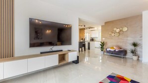 A modern living room with a large flat-screen TV, a wooden console table, a colorful rug, and a kitchen area in the background.