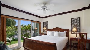 Une chambre spacieuse avec un grand lit, un bureau et une chaise. Elle est équipée d’un ventilateur de plafond et offre une vue sur un jardin grâce à une porte-fenêtre coulissante.