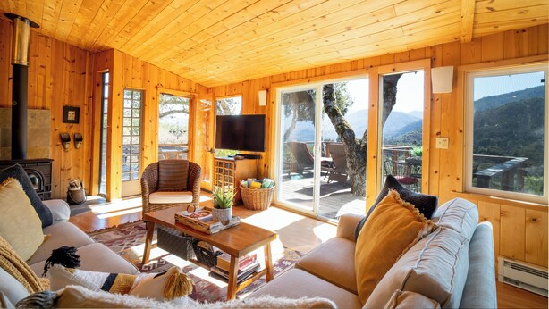 A cozy wooden cabin interior with a fireplace, comfortable seating, a flat-screen TV, and large windows offering a scenic mountain view. - Gated Carmel Valley Retreat with 20 Acres, Pool & Hot Tub (Carmel Valley)