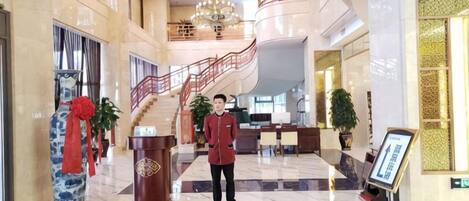 A hotel lobby with a marble floor, a reception desk, a staircase, and a large vase with a red ribbon.
