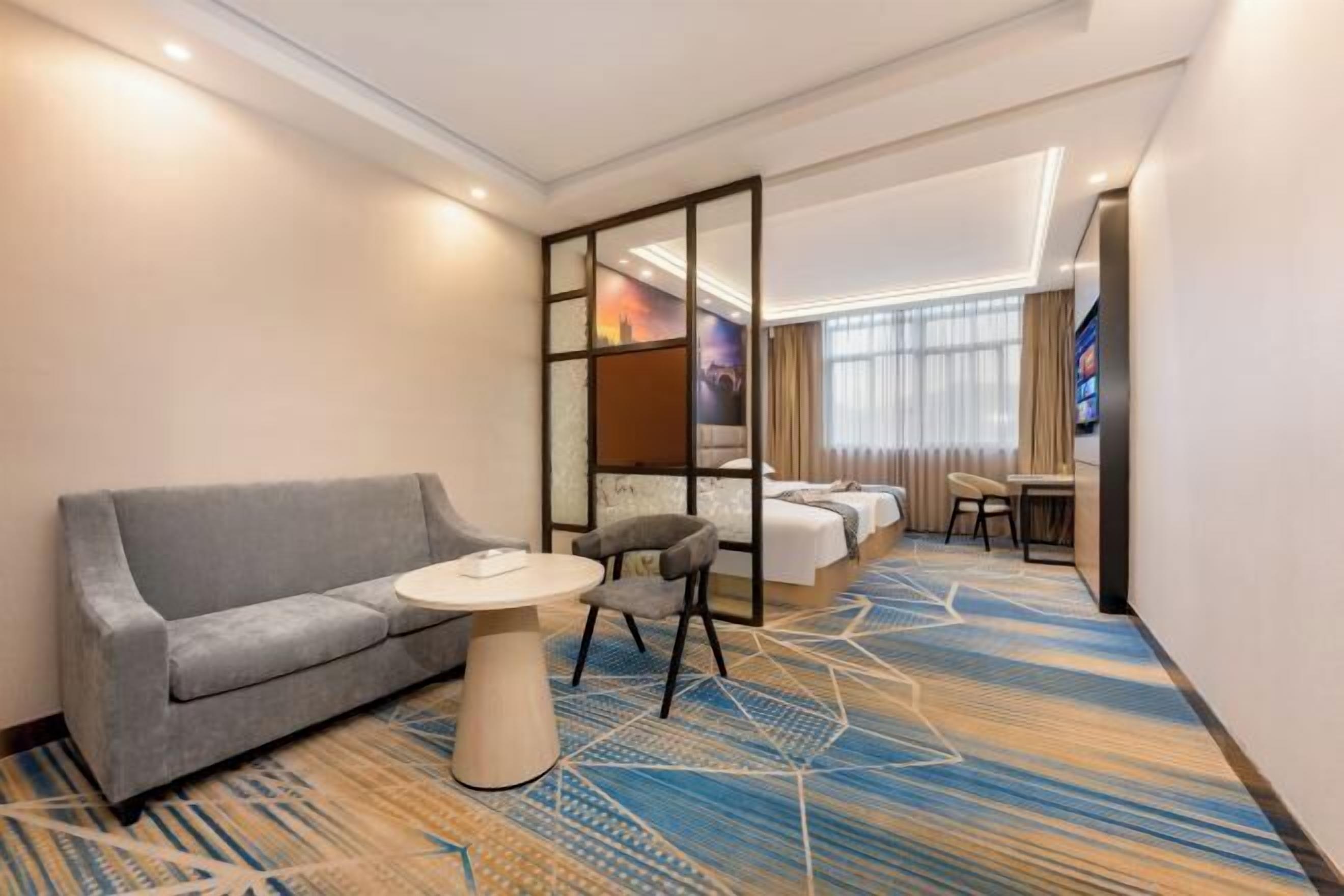 A hotel room with a sofa, a small round table, a chair, a bed, a desk, and a large window.