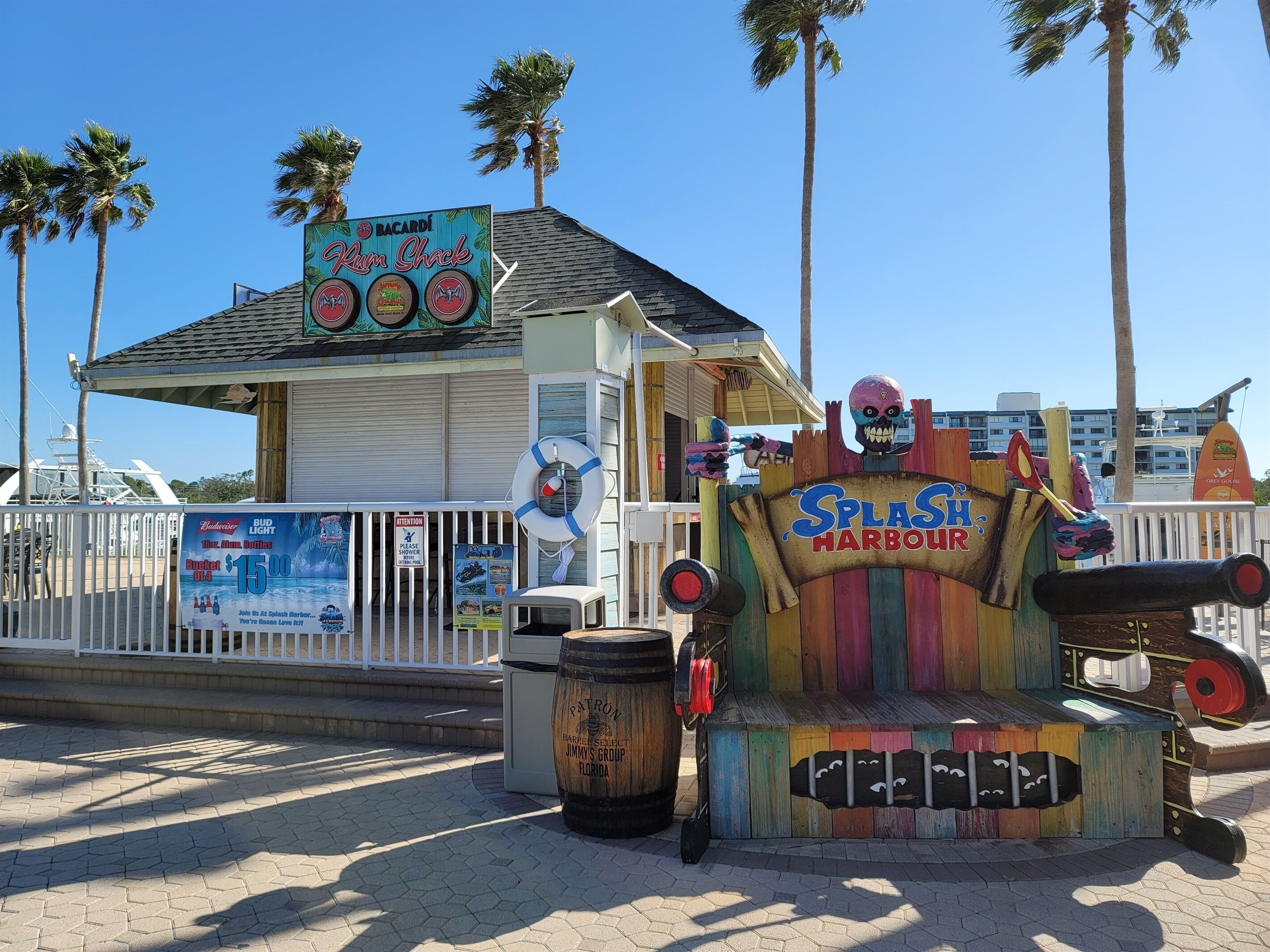 Un letrero de temática pirata y colores llamativos, con un flotador salvavidas y un barril que dice "Patron". En el fondo, un edificio muestra un letrero para "BACARDI Rum Shack" y un anuncio de Bud Light.