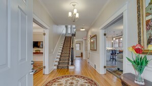 A spacious hallway with a wooden floor, a patterned rug, a staircase with a carpeted landing, and a dining area in the background. - Historic 4-Bedroom Manor | Walk to Music Row & Vanderbilt | Family Retreat (Nashville)