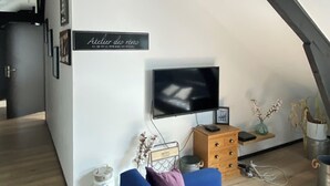 Ein gemütliches Loft-Apartment mit freiliegenden Holzbalken, einem an der Wand montierten Flachbildfernseher, einem blauen Sofa und einer kleinen Holzkabine.