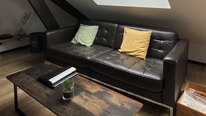 Ein Loft-Zimmer mit schrägem Dach, einem schwarzen Ledersofa mit Kissen, einem Holztisch und einem Dachfenster.